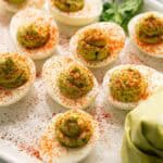 A platter of Avocado Deviled Eggs topped with green filling and sprinkled with paprika sits next to a piping bag filled with the creamy avocado mixture.