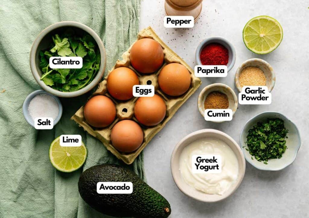 A top view of ingredients for Avocado Deviled Eggs, including eggs, avocado, cilantro, Greek yogurt, lime, salt, pepper, paprika, cumin, and garlic powder arranged on a white surface.