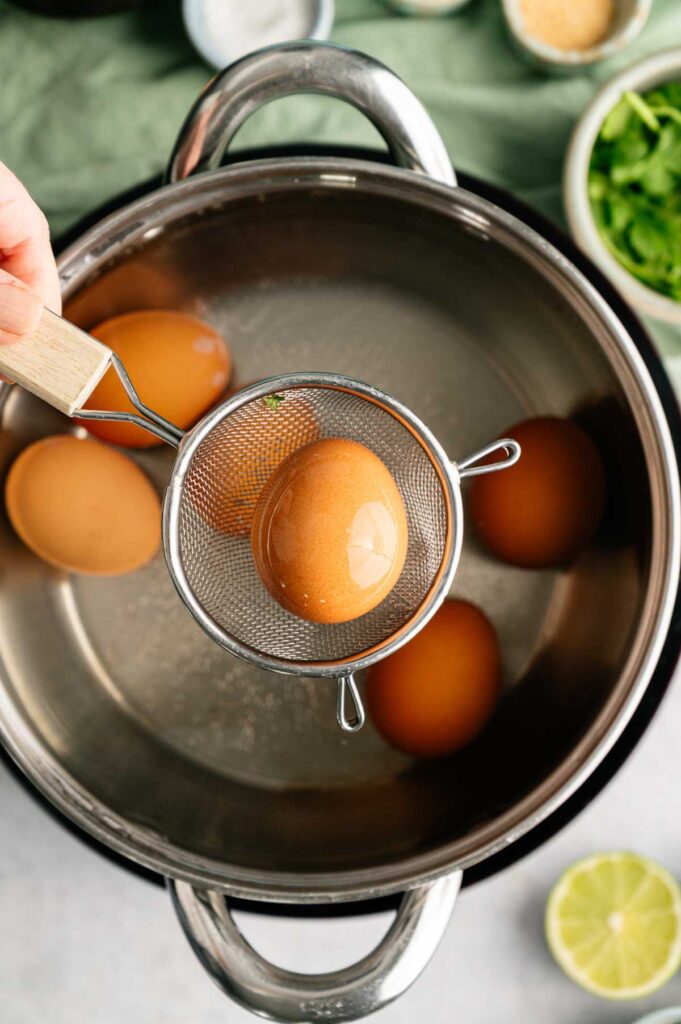 A hand holds a mesh strainer lifting a brown egg from a pot of water with three other eggs inside, preparing the perfect base for Avocado Deviled Eggs. Bowls of salt, greens, and a lime are nearby, ready to elevate the flavor of this delicious dish.