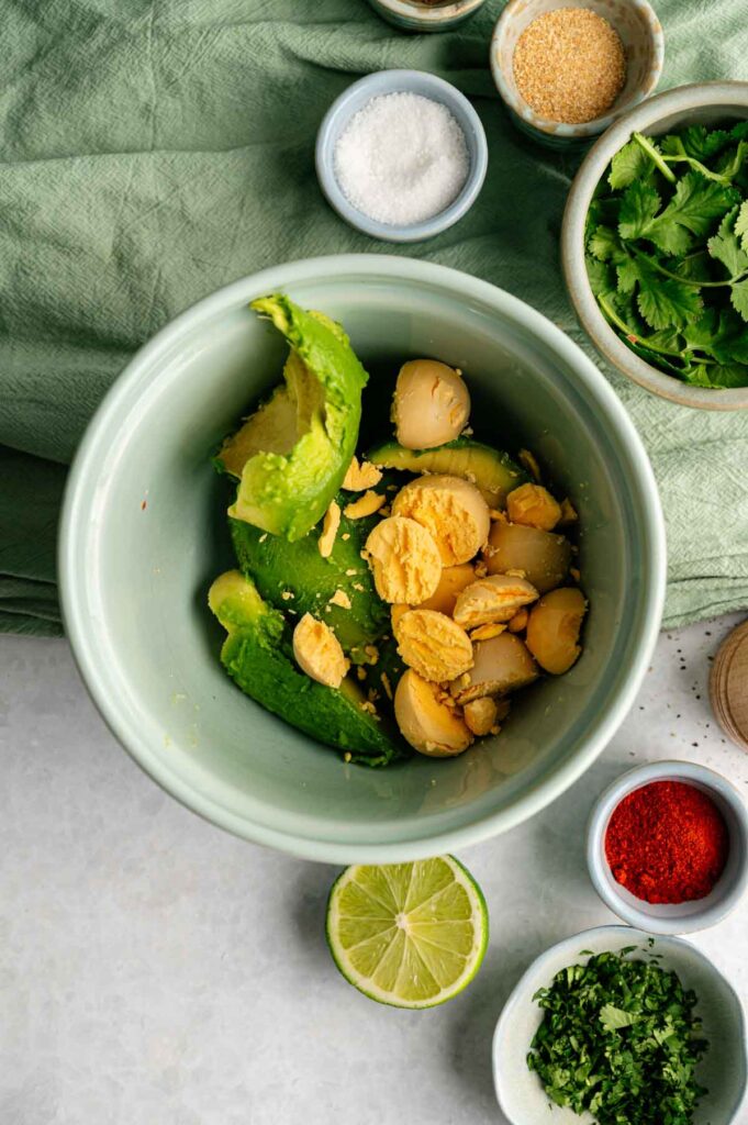 A bowl with avocado and hard-boiled egg yolks, ready to become delicious Avocado Deviled Eggs, surrounded by fresh herbs, lime, salt, and spices on a countertop.