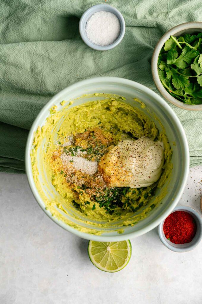 A bowl with mashed avocado, mayonnaise, spices, fresh herbs, and salt—perfect ingredients for Avocado Deviled Eggs—is surrounded by cilantro, a lime half, red spice, and a green cloth.