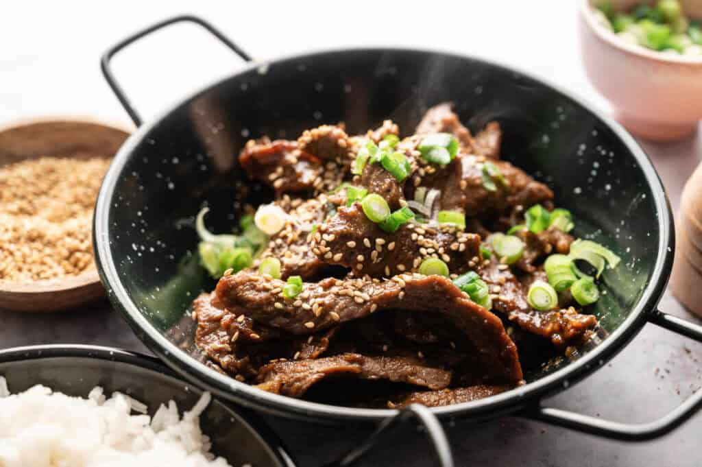 A black bowl filled with sliced cooked Beef Bulgogi topped with sesame seeds and chopped green onions, served alongside a bowl of rice and extra sesame seeds.