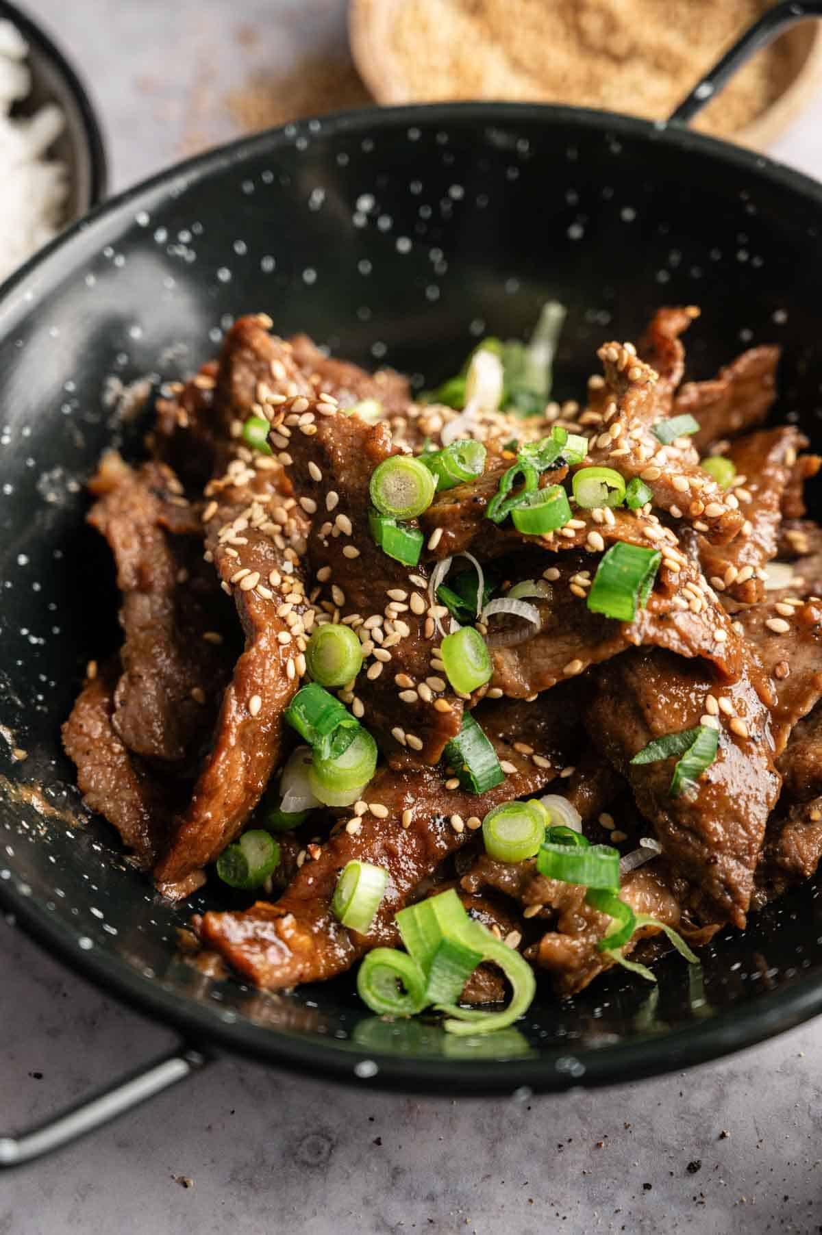 A black bowl filled with sliced Beef Bulgogi, topped with sesame seeds and chopped green onions, sits on a gray surface.