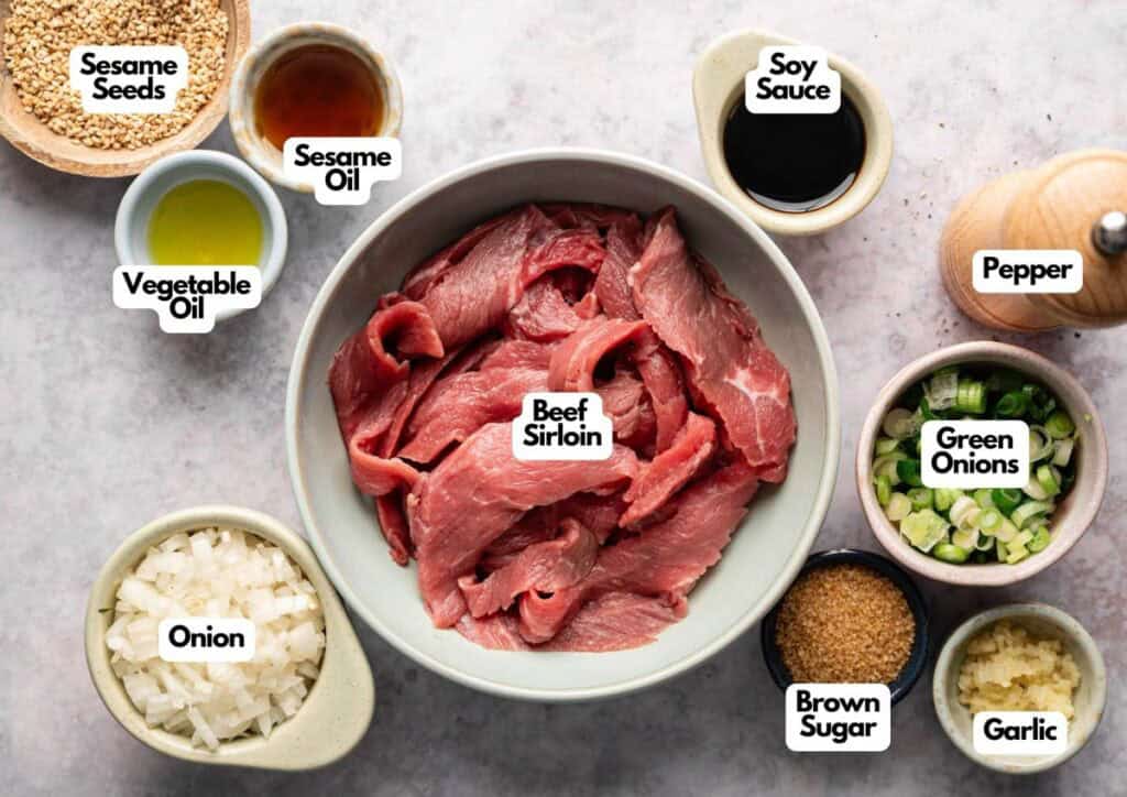 Sliced beef sirloin in a bowl, surrounded by small bowls of diced onion, green onions, brown sugar, garlic, pepper, soy sauce, vegetable oil, sesame oil, and sesame seeds—perfect ingredients for making authentic Beef Bulgogi.