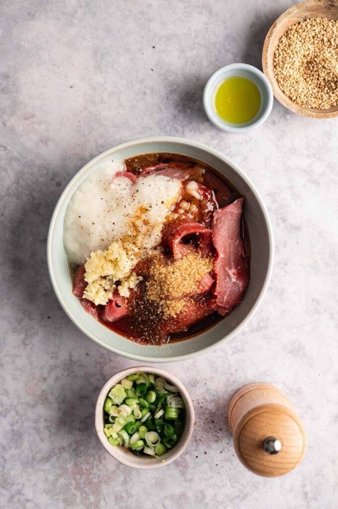 A bowl with sliced beef, grated onion, minced garlic, brown sugar, soy sauce, and spices—perfect for making Beef Bulgogi—sits on a countertop near green onions, sesame seeds, olive oil, and a pepper grinder.