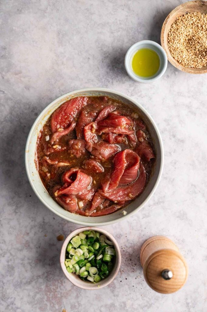A bowl of marinated raw beef bulgogi sits on a countertop, surrounded by chopped green onions, olive oil, sesame seeds, and a pepper grinder.