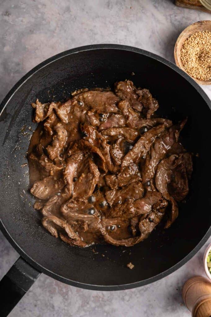 Sliced beef bulgogi cooking in a black skillet with sauce, seen from above, on a gray countertop.