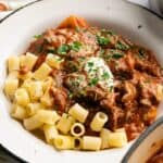A plate of pasta with a hearty goulash beef stew in tomato sauce, topped with a dollop of sour cream and chopped parsley.