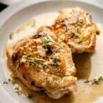 Two pieces of Tuscan Chicken Thighs with golden-brown skin served in a creamy sauce, garnished with fresh rosemary on a white plate.