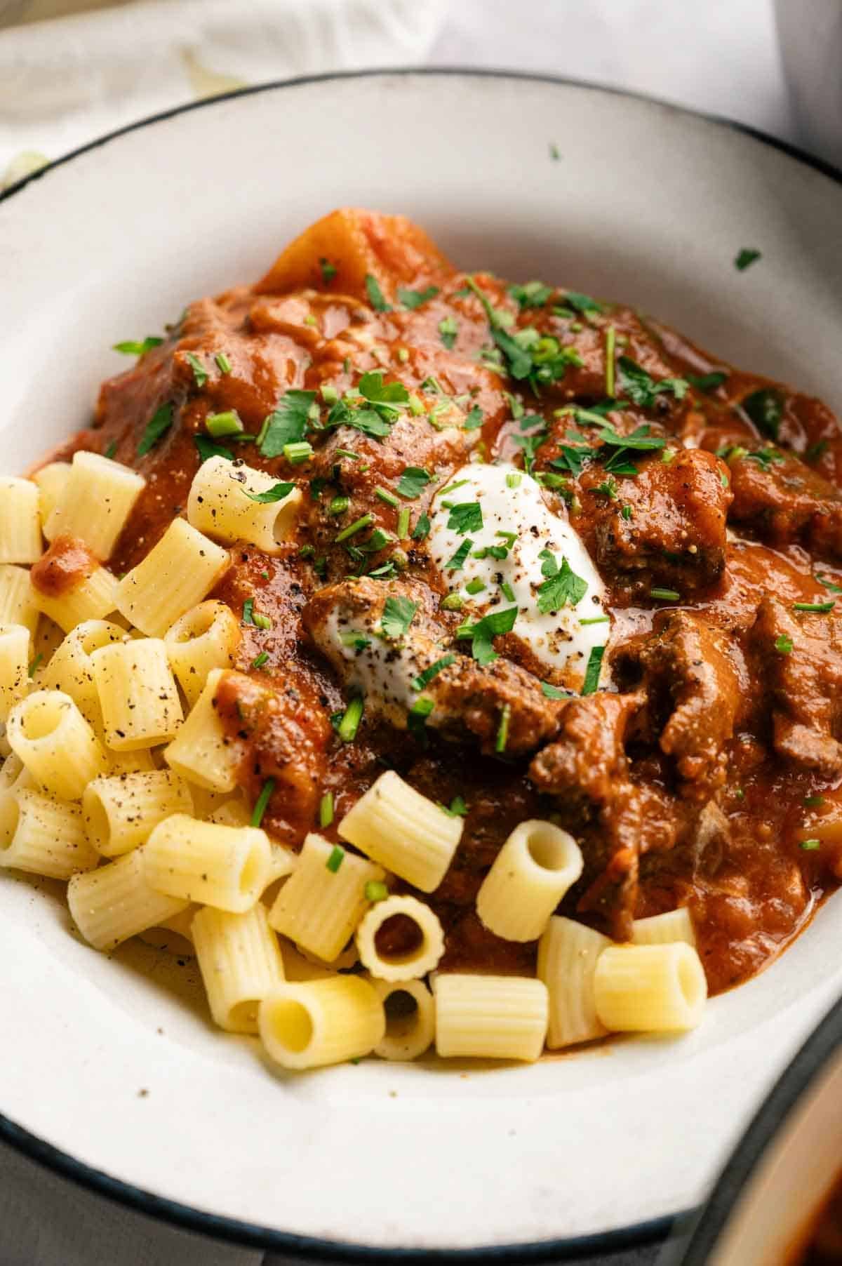 A plate of pasta topped with hearty goulash beef stew in tomato sauce, garnished with a dollop of sour cream and chopped herbs.