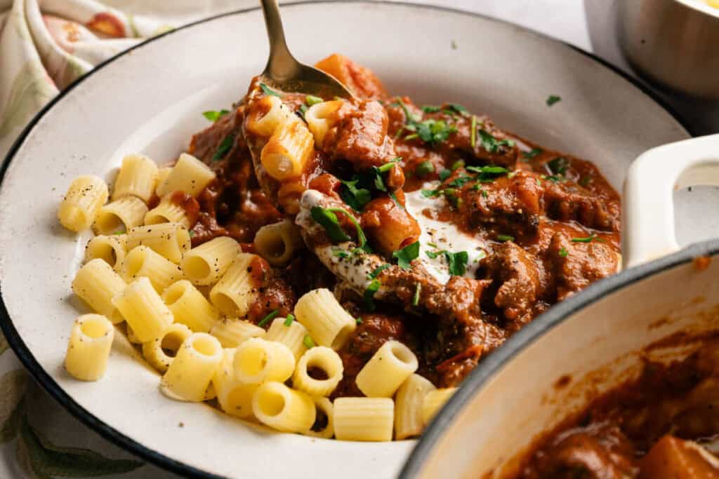A plate of cooked pasta sits next to a hearty serving of beef goulash in tomato sauce, garnished with chopped parsley, as a fork scoops up a flavorful bite.