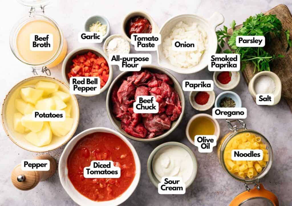Various labeled ingredients for hearty goulash or beef stew, including beef chuck, potatoes, diced tomatoes, bell pepper, onion, spices, broth, noodles, sour cream, parsley, and olive oil arranged on a table.