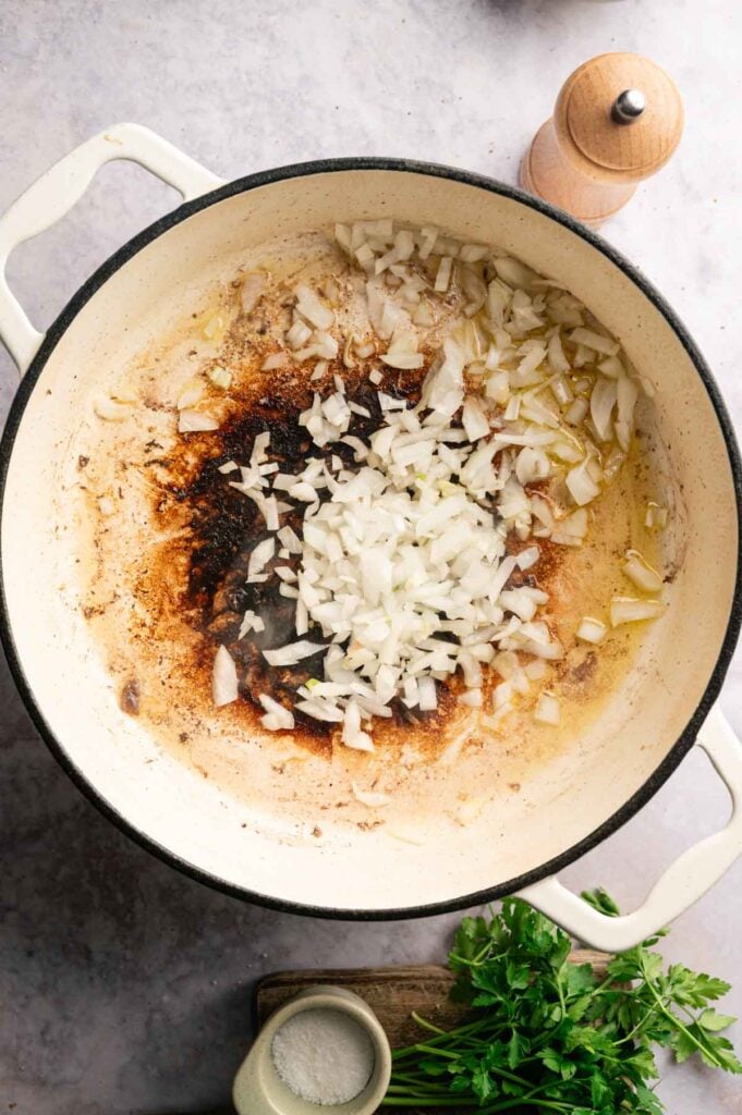 Chopped onions and oil are sautéing in a white pot with browned bits on the bottom, creating a flavorful base for goulash, next to a pepper grinder, salt, and fresh parsley.