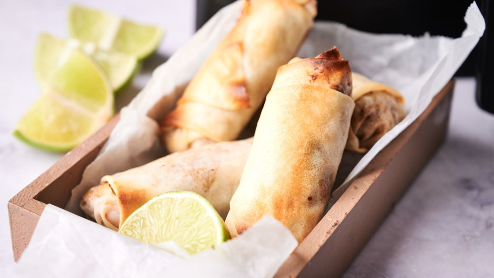 Three golden-brown spring rolls in a wooden tray lined with parchment paper, served with lime wedges.
