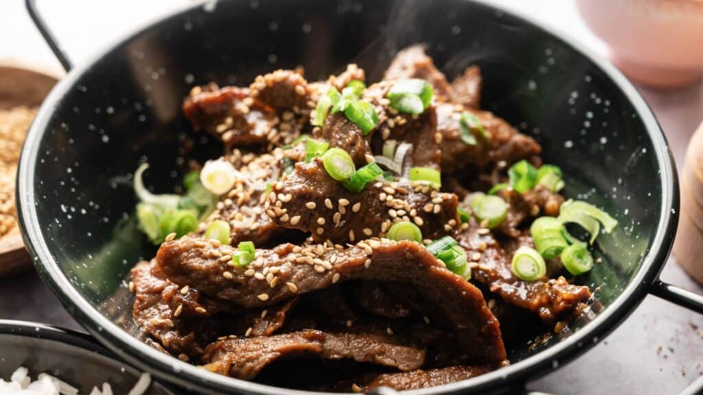 A bowl of sliced beef topped with sesame seeds and chopped green onions, served in a black dish.