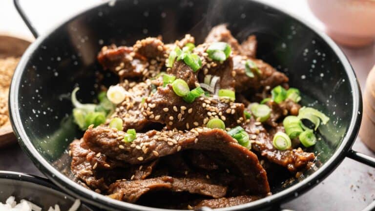 A bowl of sliced beef topped with sesame seeds and chopped green onions, served in a black dish.
