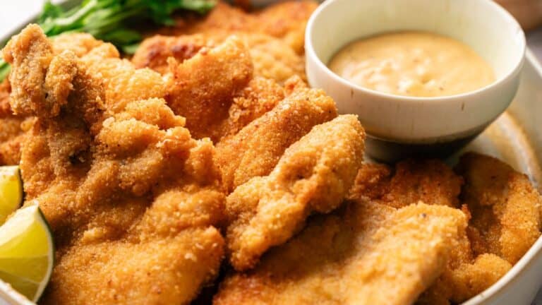 A plate of breaded and fried chicken cutlets with a dipping sauce in a bowl, garnished with lime wedges and green herbs.