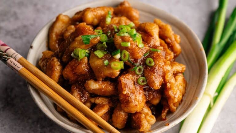 A bowl of glazed chicken pieces topped with chopped green onions, with a pair of chopsticks resting on the rim, and green onions beside the bowl.