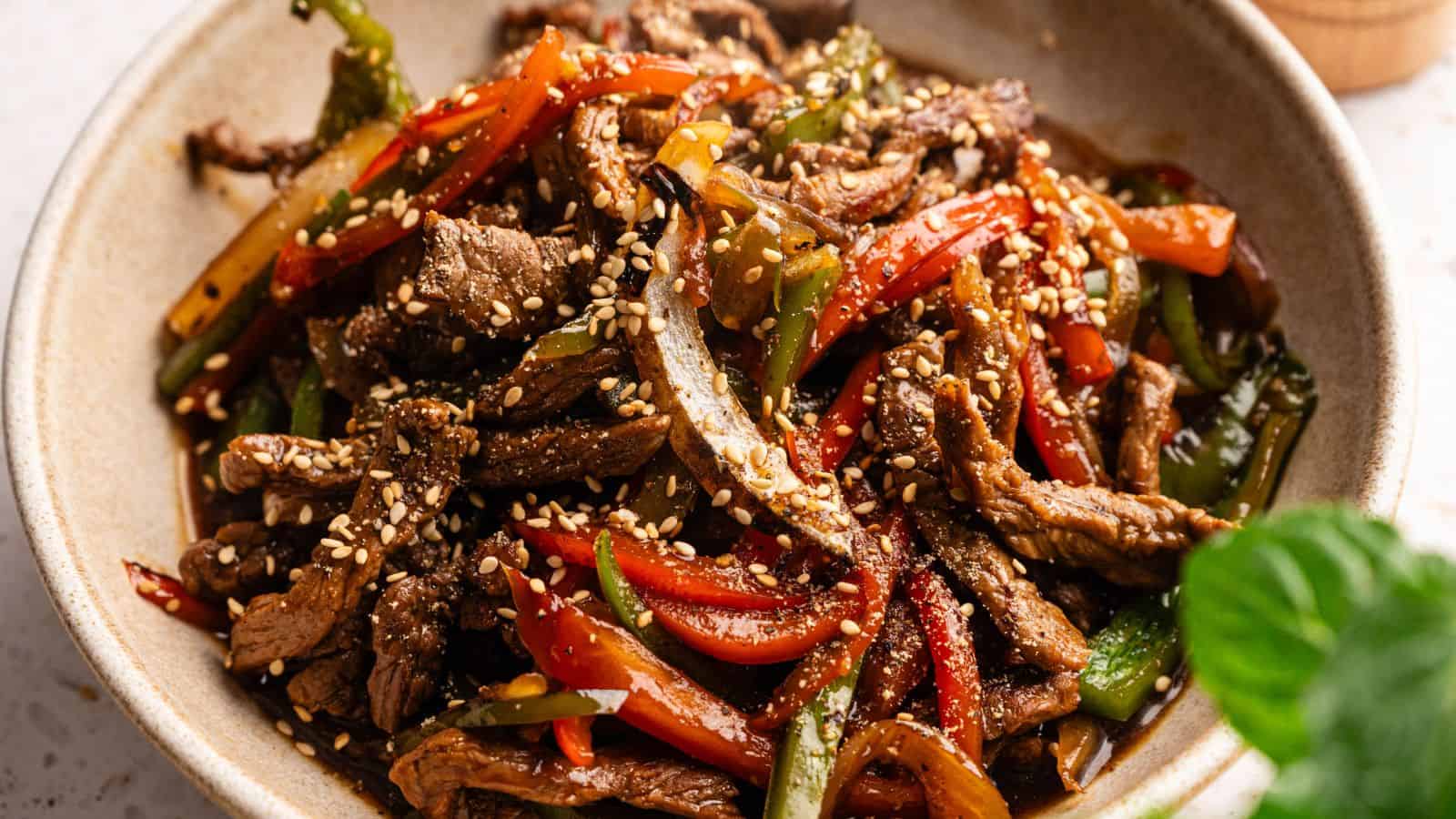 A bowl of stir-fried beef strips with red and green bell peppers, topped with sesame seeds.