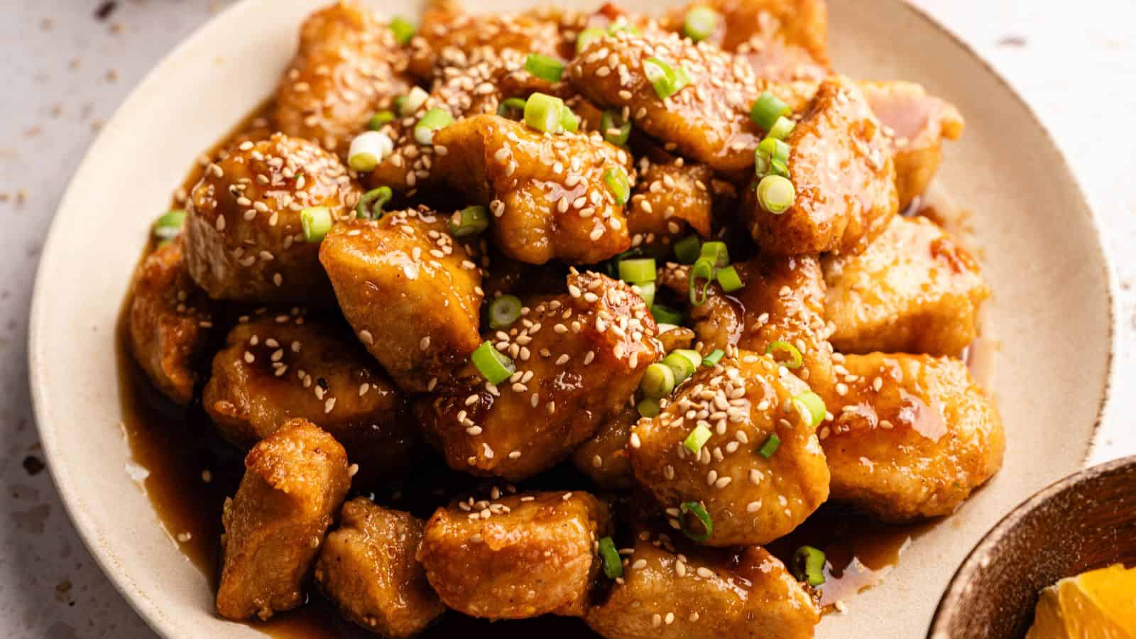 A plate of sesame chicken pieces covered in sauce, garnished with sesame seeds and chopped green onions.