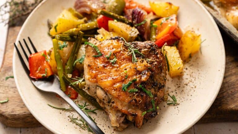 A plate with roasted chicken thigh, assorted roasted vegetables including potatoes, red peppers, green beans, and onions, garnished with herbs. A black fork rests on the plate.