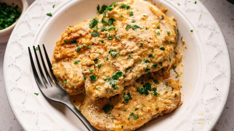 Three pieces of creamy, herb-garnished chicken are served on a white plate with a black fork resting beside them.
