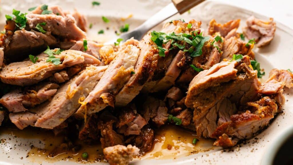 Sliced roasted pork garnished with chopped herbs, served on a white plate with visible juices and seasoning.