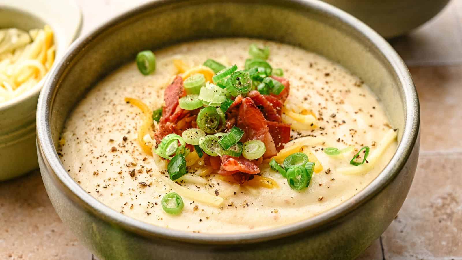 A bowl of creamy soup topped with chopped green onions, grated cheese, crispy bacon pieces, and ground black pepper.