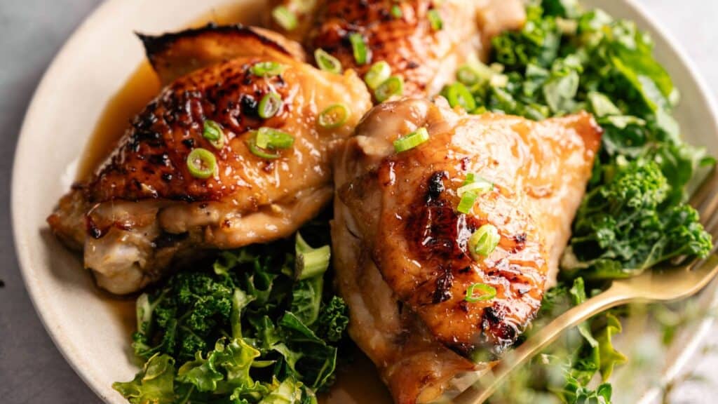 Three pieces of glazed roasted chicken sit on a bed of leafy greens, garnished with chopped green onions on a beige plate.
