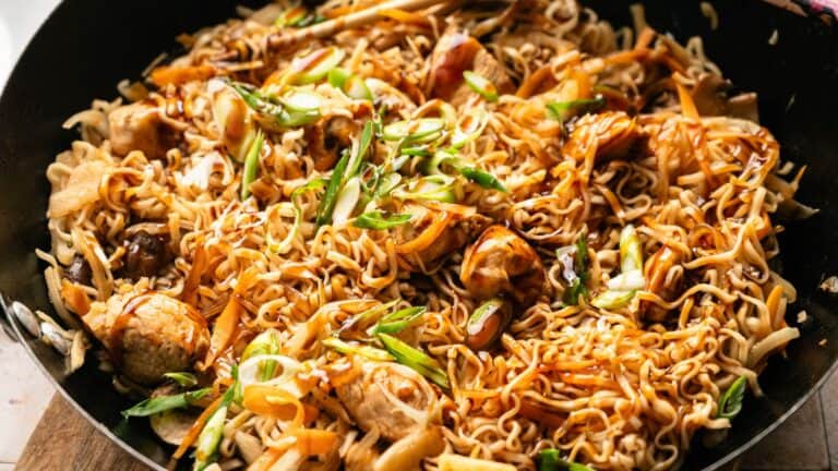 A pan filled with cooked noodles, vegetables, chicken pieces, and garnished with sliced green onions in a savory sauce.