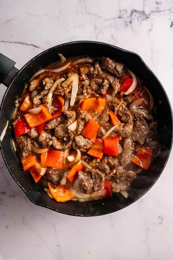 A skillet on a marble surface filled with cooked beef strips, red bell peppers, and sliced onions in a rich brown sauce, reminiscent of Panda Express Beijing Beef.