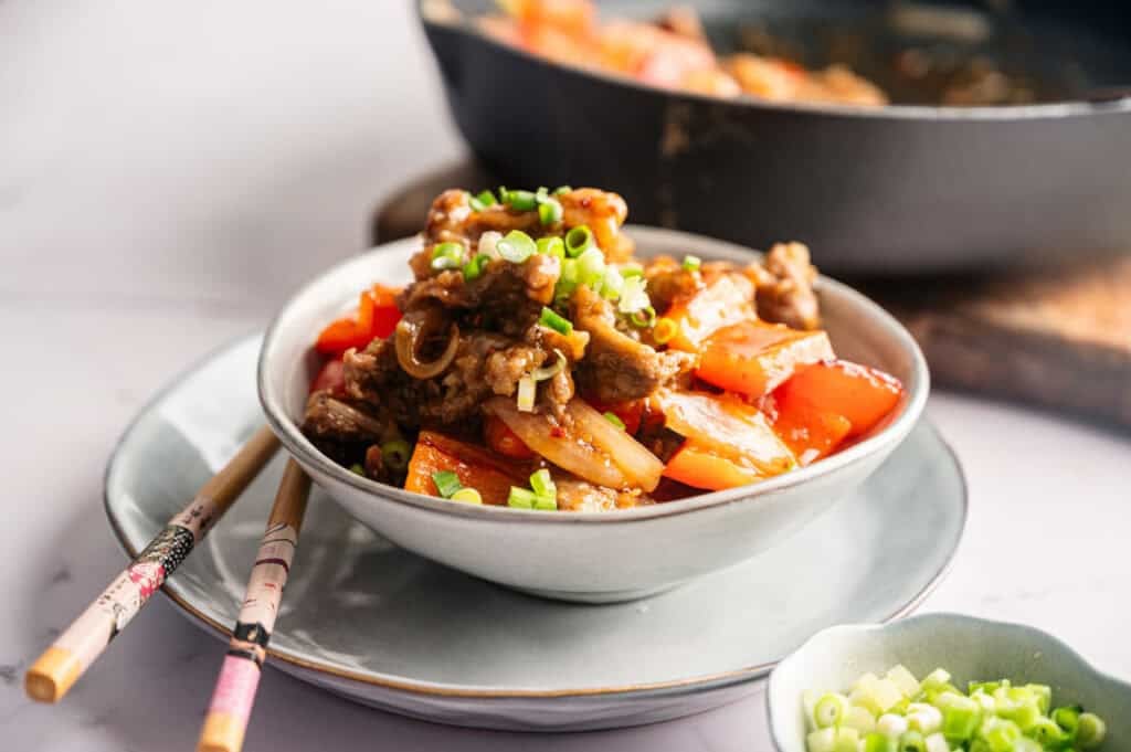 A bowl of stir-fried beef with tomatoes and green onions, inspired by Panda Express Beijing Beef, served with chopsticks on a plate, with more food and a small dish of chopped green onions in the background.