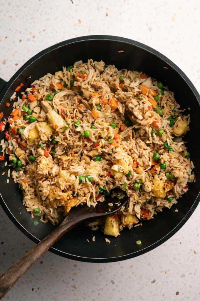 A black pan filled with Pineapple Fried Rice containing peas, carrots, pineapple chunks, and shredded chicken, with a wooden spoon resting inside.
