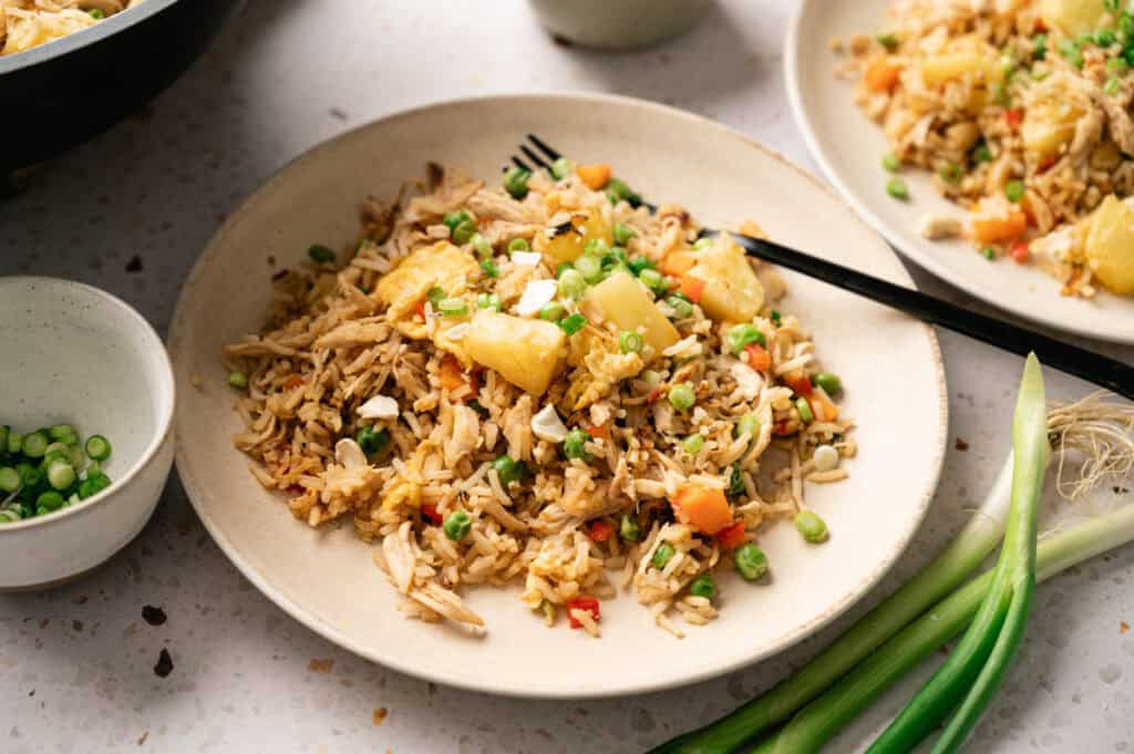 A flavorful plate of Pineapple Fried Rice with shredded chicken, peas, carrots, chopped green onions, and cashews, served with chopsticks and extra green onions on the side.