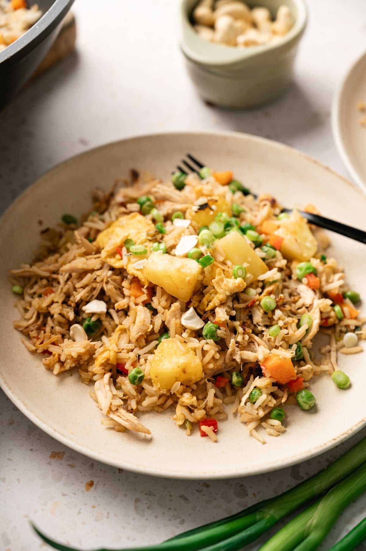 A plate of Pineapple Fried Rice features pineapple chunks, green peas, diced carrots, shredded chicken, and chopped cashews, with a black fork on the side. Green beans and a bowl of cashews are nearby.