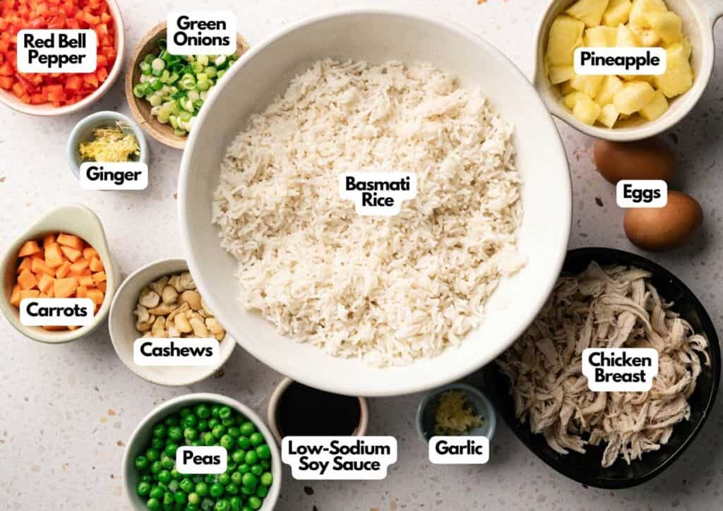 A bowl of Pineapple Fried Rice surrounded by labeled ingredients: red bell pepper, green onions, pineapple, eggs, chicken breast, garlic, low-sodium soy sauce, peas, cashews, carrots, and ginger.