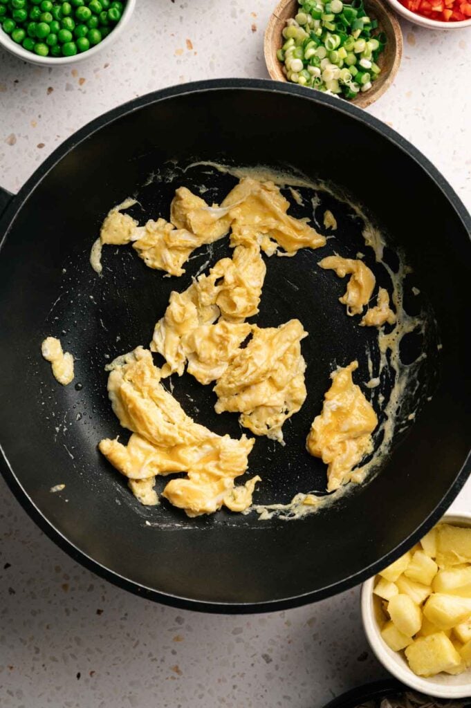 Softly scrambled eggs cooking in a black skillet, with pineapple and peas nearby.