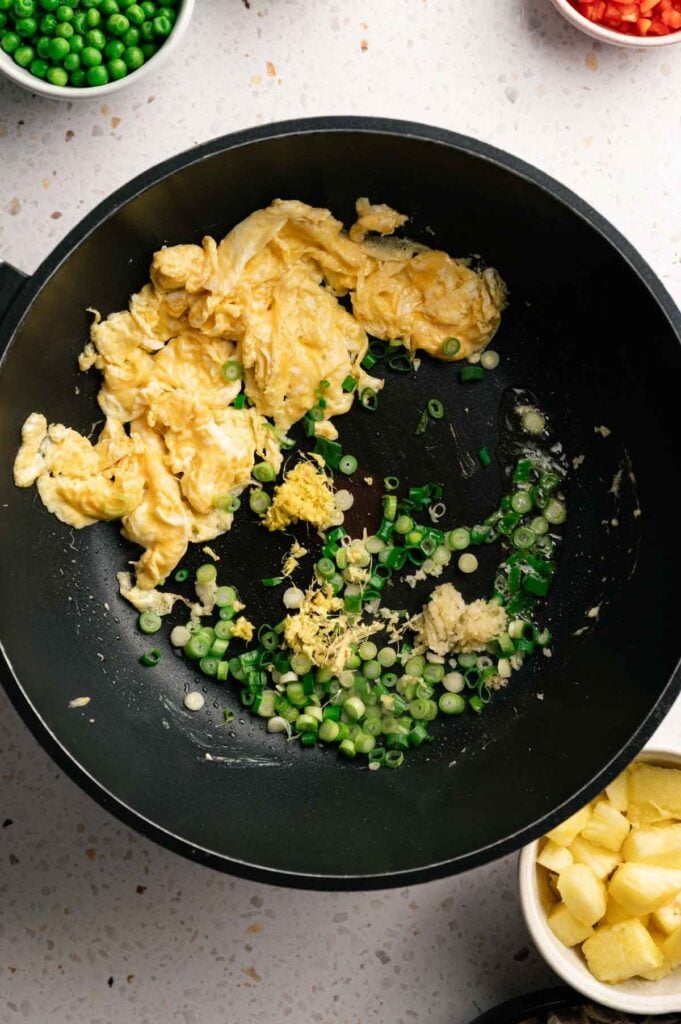 Scrambled eggs pushed aside with green onion, garlic, and ginger sautéing in a wok.