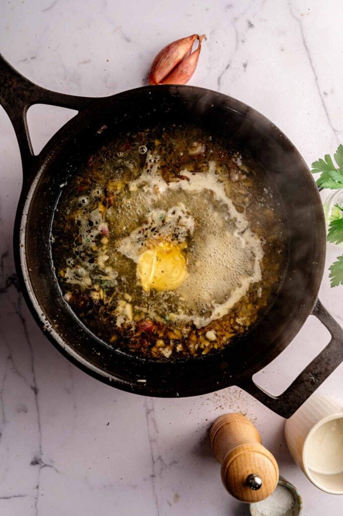 A cast iron pan with melted butter, chopped onions, and herbs sizzles on a marble countertop—the perfect start for Creamy Skillet Chicken Thighs. Nearby, a shallot, pepper grinder, parsley, and cream await to add flavor and richness to this comforting dish.