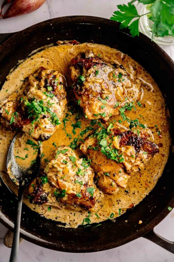 Four pieces of Creamy Skillet Chicken Thighs in a luscious sauce, garnished with chopped parsley, served in a black skillet with a spoon.