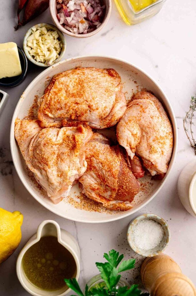 Raw chicken thighs seasoned with spices in a bowl, surrounded by chopped garlic, shallots, butter, oil, lemon, salt, pepper, and fresh herbs on a marble countertop—perfect for preparing Creamy Skillet Chicken Thighs.