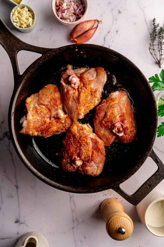 Four seasoned raw chicken thighs in a cast iron skillet, surrounded by bowls of chopped garlic, shallots, herbs, a pepper grinder, and a jug on a marble countertop—perfectly prepped for making delicious Creamy Skillet Chicken Thighs.