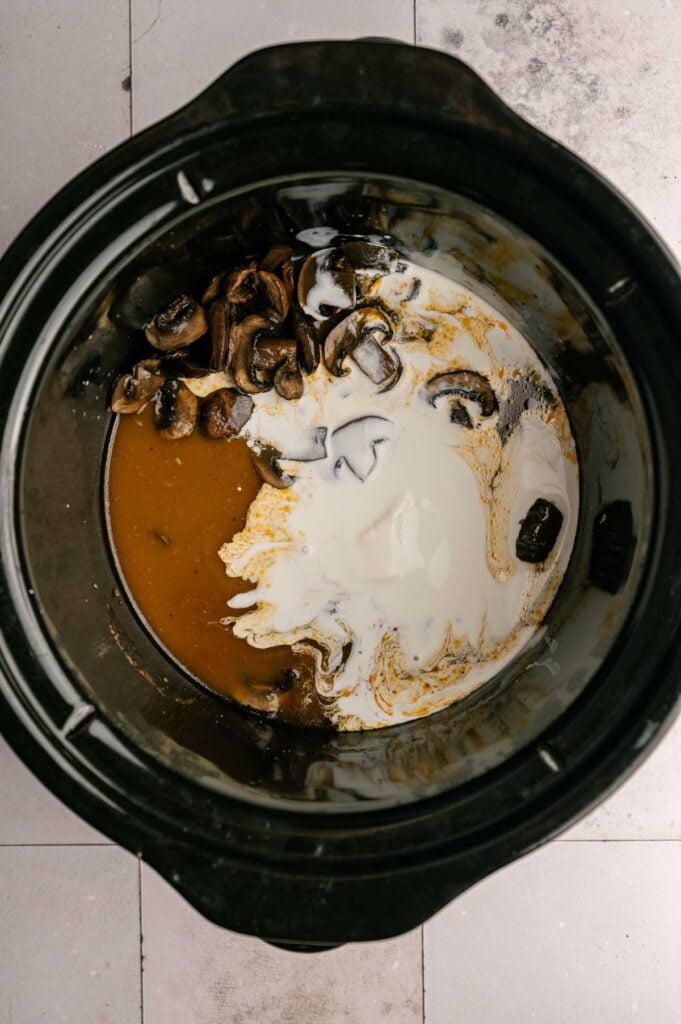 Overhead view of a slow cooker with sliced mushrooms, brown broth, and cream being mixed together on a tiled surface—a delicious base for tender Slow Cooker Chicken Breast.