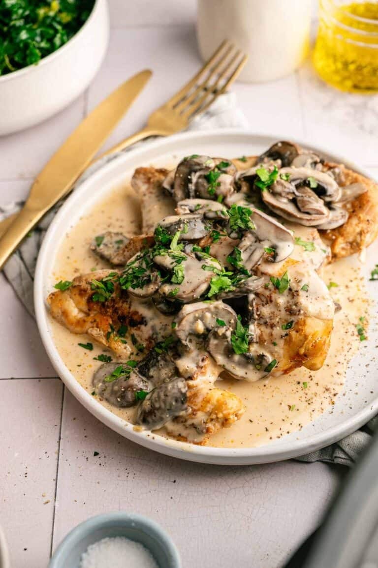 Plate of slow cooker chicken breast topped with creamy mushroom sauce and chopped parsley, served with a fork and knife on the side.
