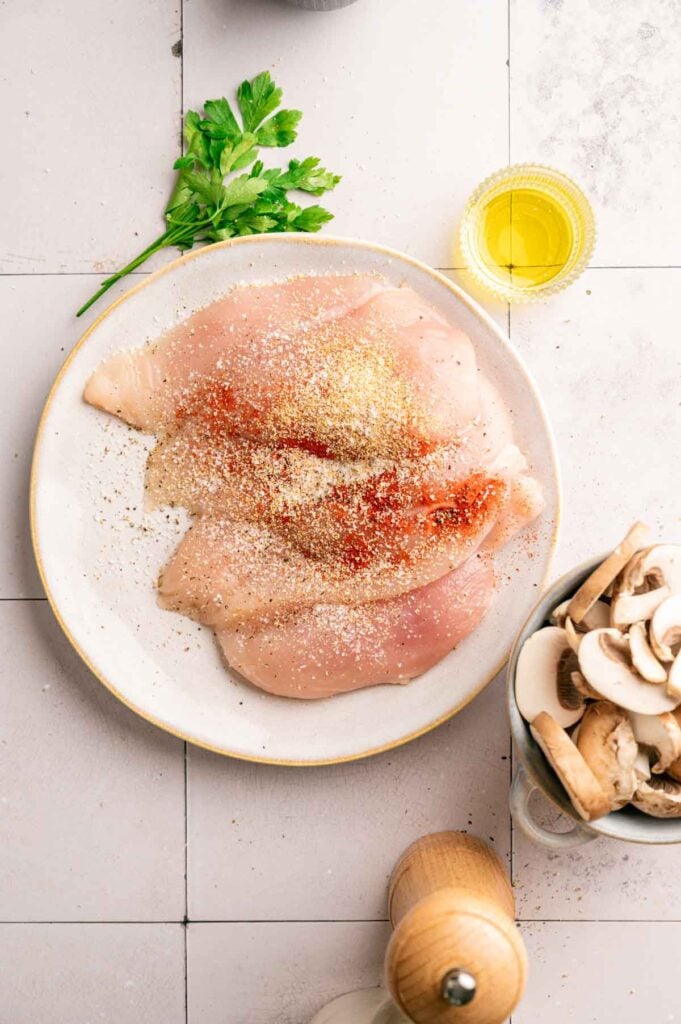 Raw chicken breasts seasoned with paprika and garlic powder on a plate, next to sliced mushrooms.