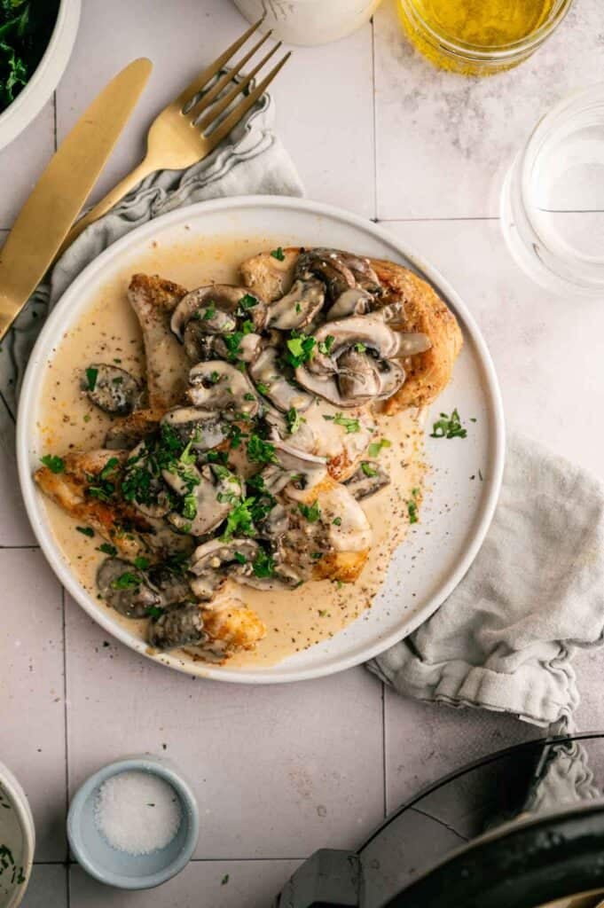 A plate of Slow Cooker Chicken Breast covered in creamy mushroom sauce, garnished with chopped herbs, sits next to a gold fork, knife, and a folded napkin.