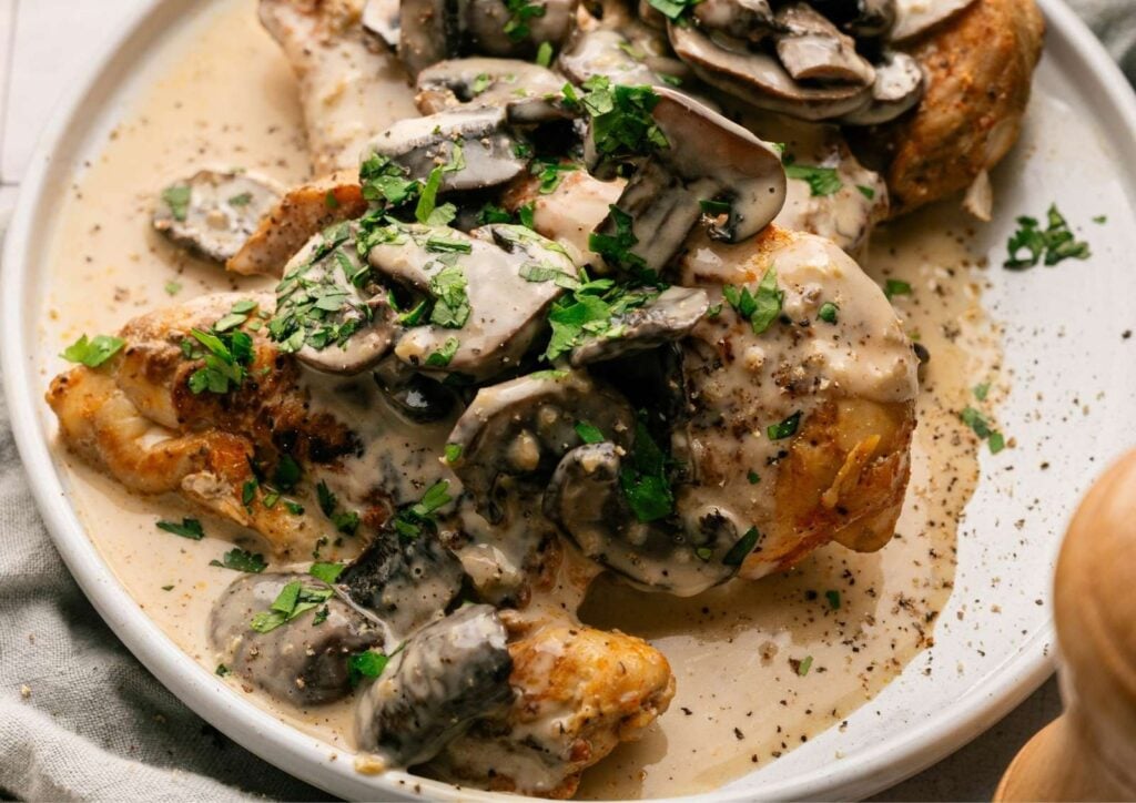 Slow Cooker Chicken Breast topped with creamy mushroom sauce and garnished with chopped parsley on a white plate.