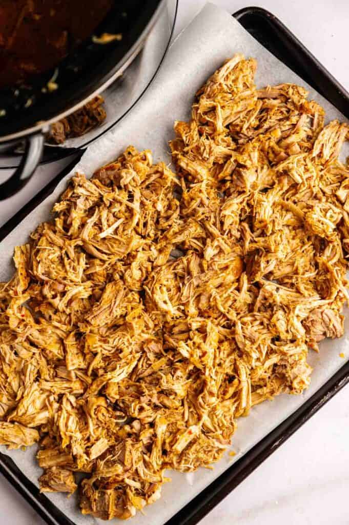 Shredded cooked chicken spread on a parchment-lined baking sheet next to a slow cooker with broth&mdash;just like prepping Slow Cooker Pork Carnitas for flavorful, tender results.