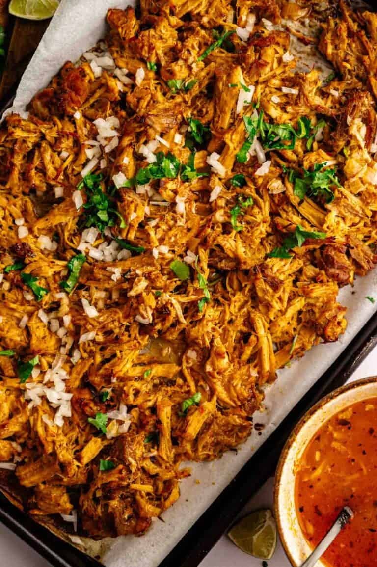 Slow Cooker Pork Carnitas shredded and garnished with chopped onion and cilantro on a baking tray, served with a bowl of reddish sauce and fresh lime wedges nearby.