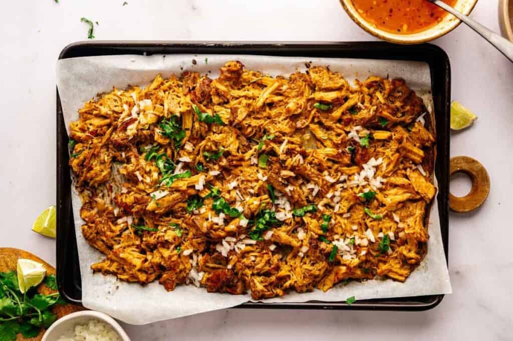 Slow Cooker Pork Carnitas shredded and topped with chopped onions and cilantro, served on a parchment-lined baking sheet with lime wedges and sauce on the side.